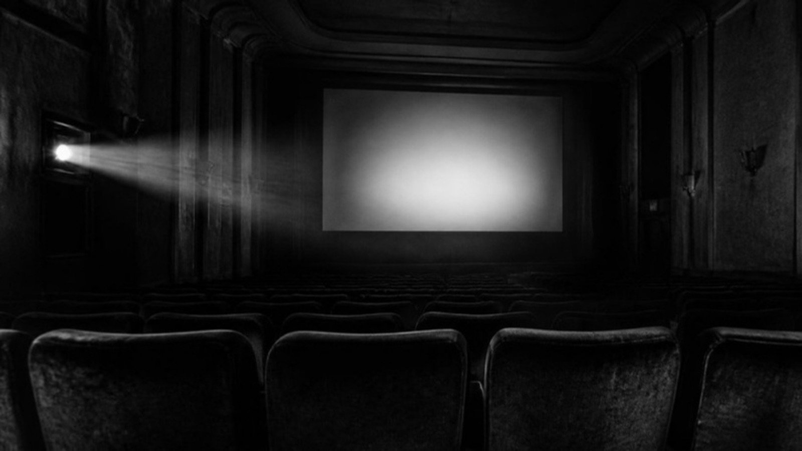 An empty old cinema hall seen from the back row: rows of seats, a blank lit screen and a projector beam through the haze.