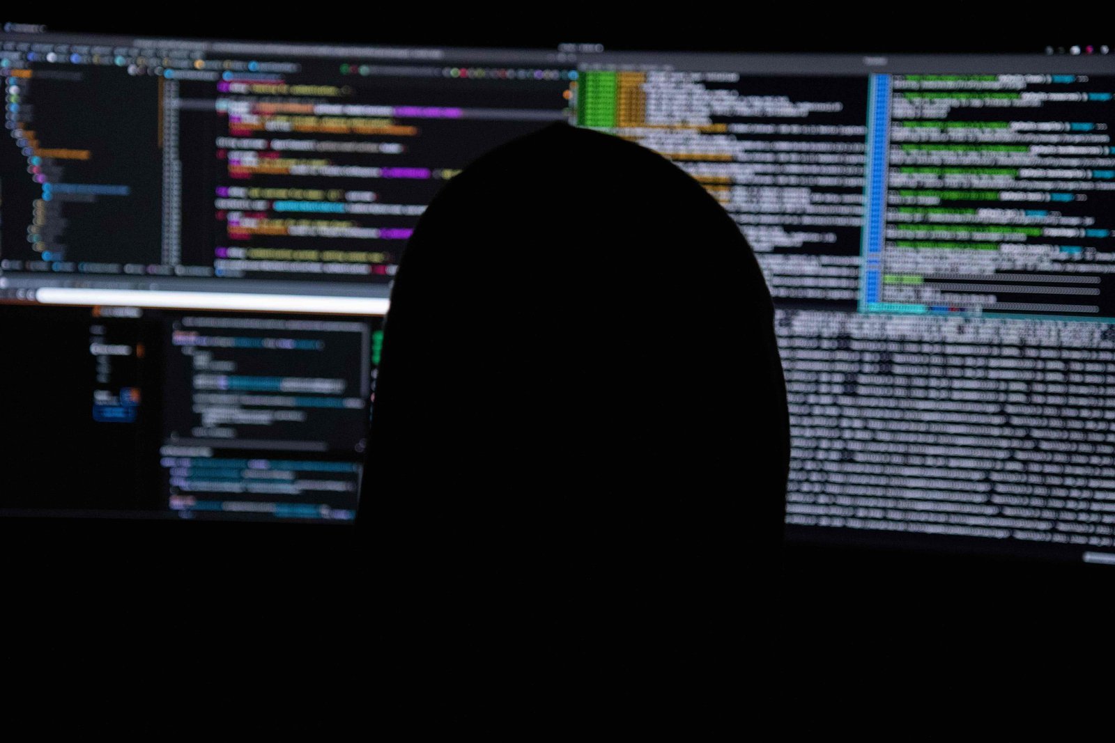 A figure in silhouette, seen from behind, facing a wall of screens filled with data.